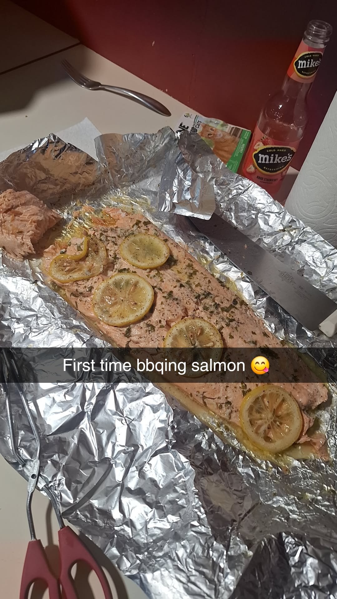 A cooked salmon fillet with lemon slices on foil, next to a knife, scissors, and a bottle of Mike’s Hard Lemonade.
