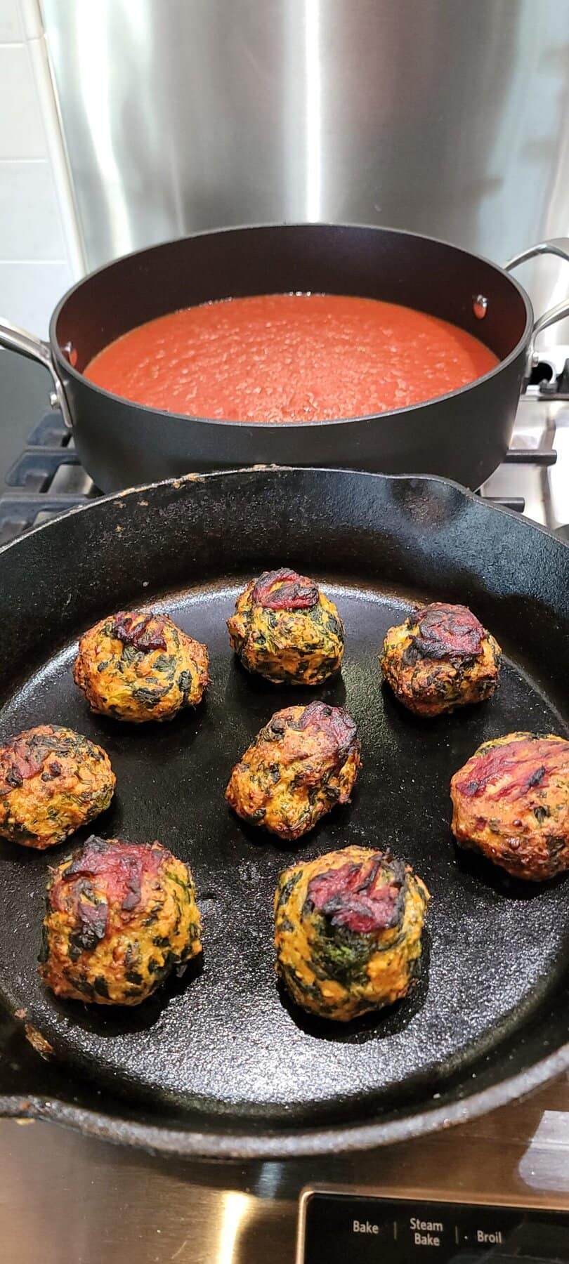 Eight baked veggie balls in a cast iron pan, with a pot of red tomato sauce simmering on the stove behind.