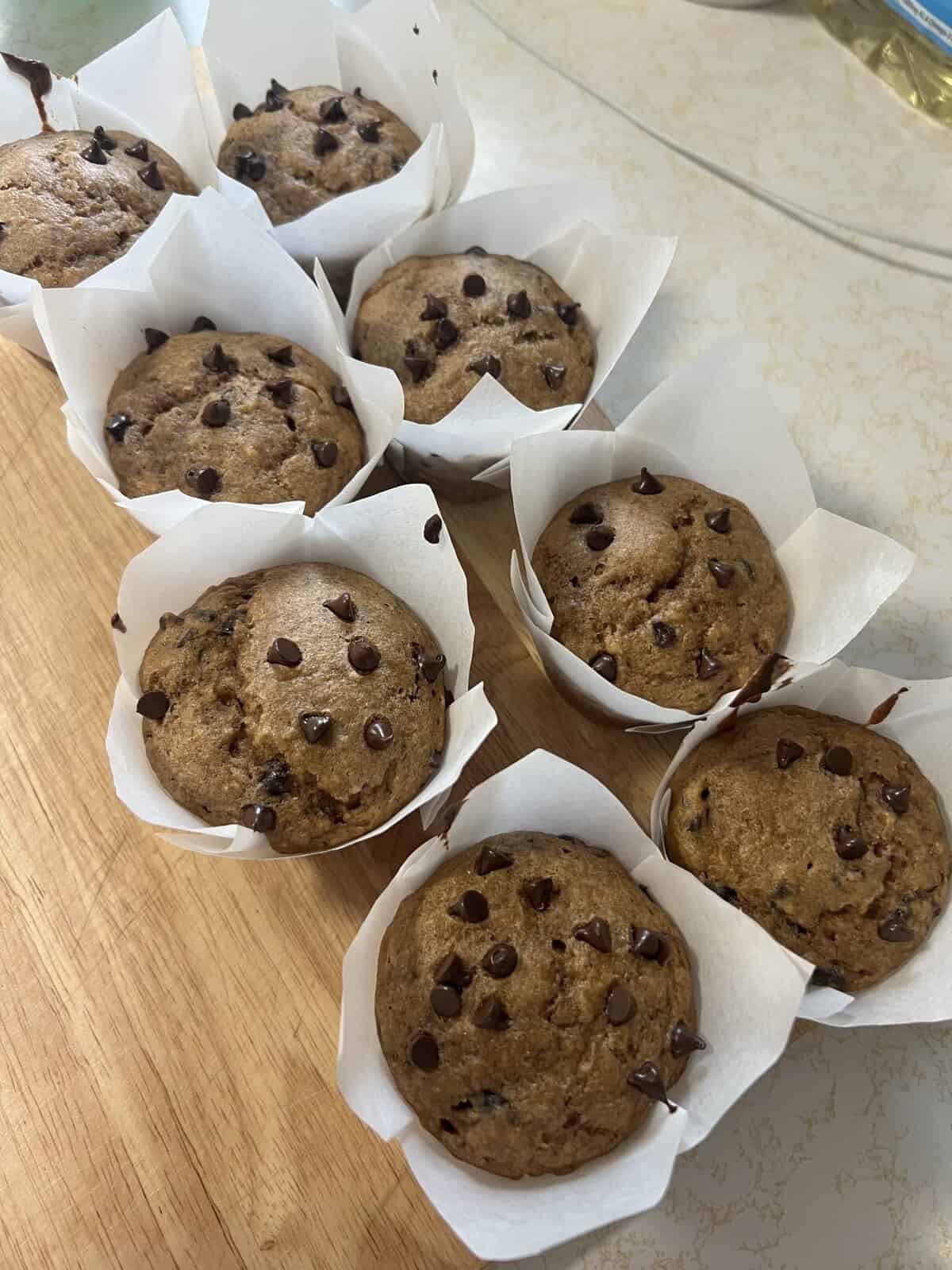 Nine chocolate chip muffins in white paper liners are arranged on a wooden cutting board.