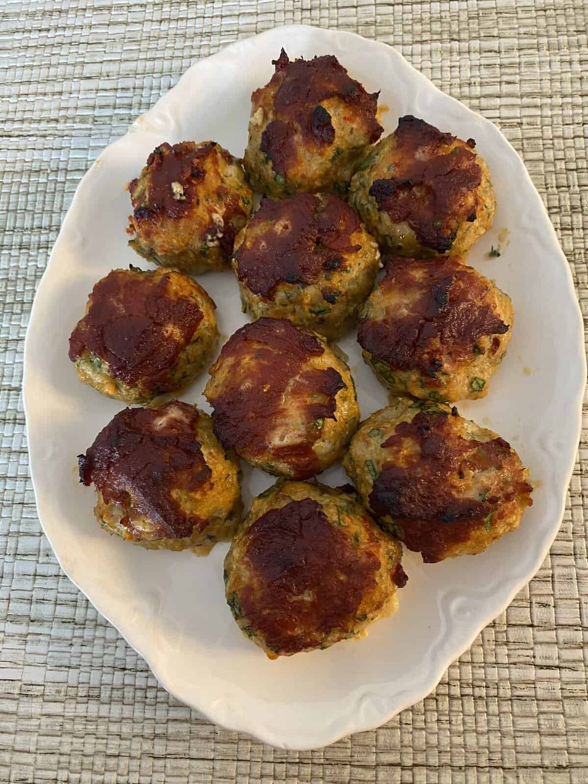 Ten browned meatballs topped with sauce are arranged on a white oval plate on a woven placemat.