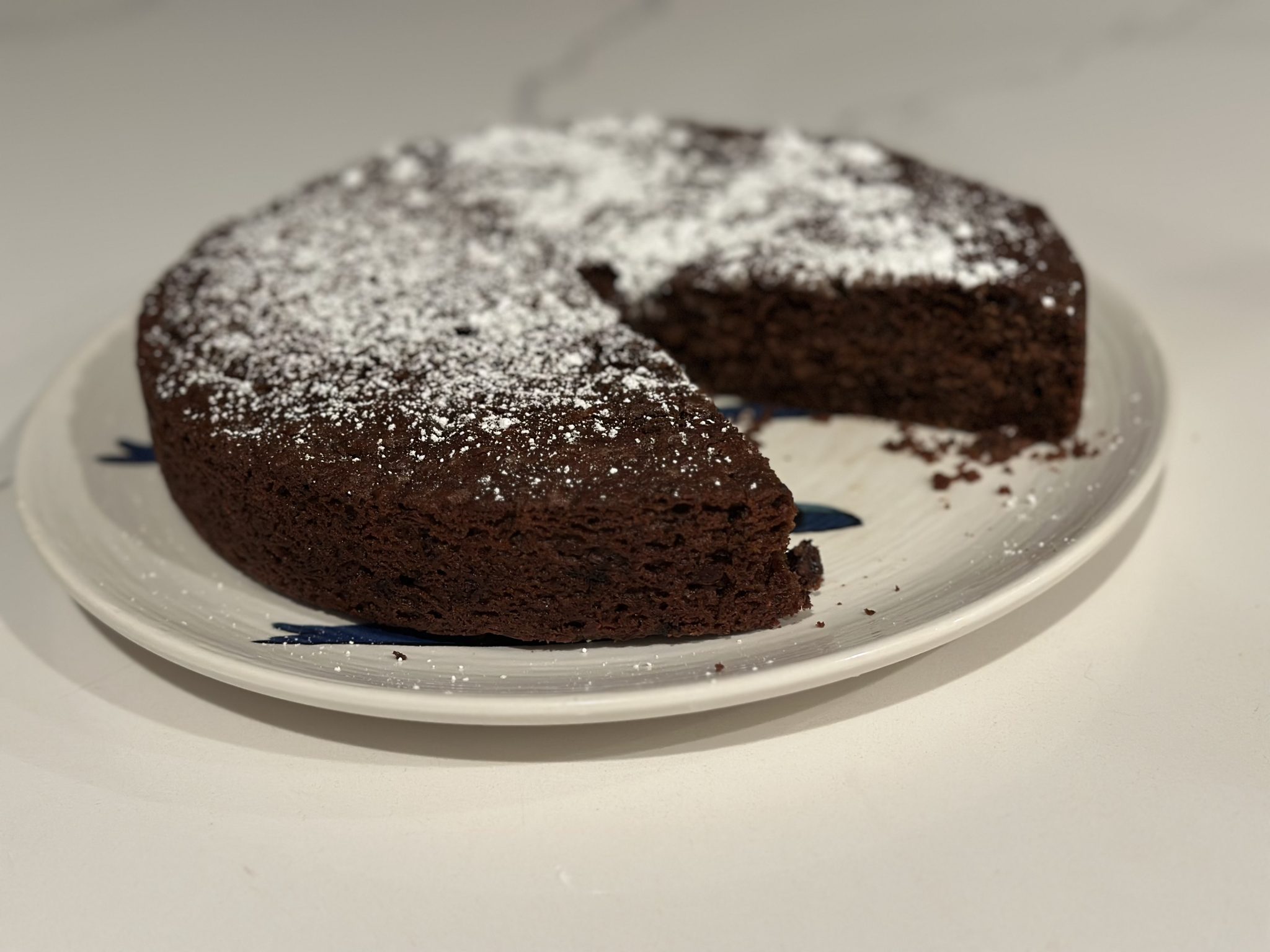 A round chocolate cake with powdered sugar on top, missing one slice, sits on a white plate.