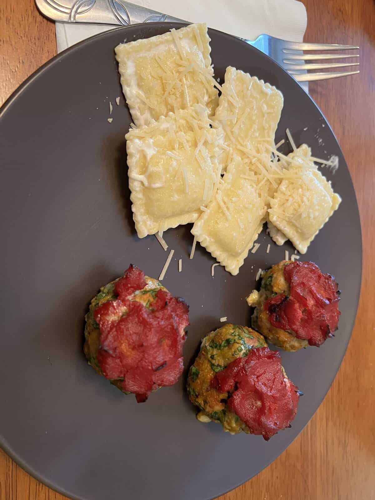 A plate with cheese ravioli topped with grated cheese and three spinach meatballs with tomato topping.