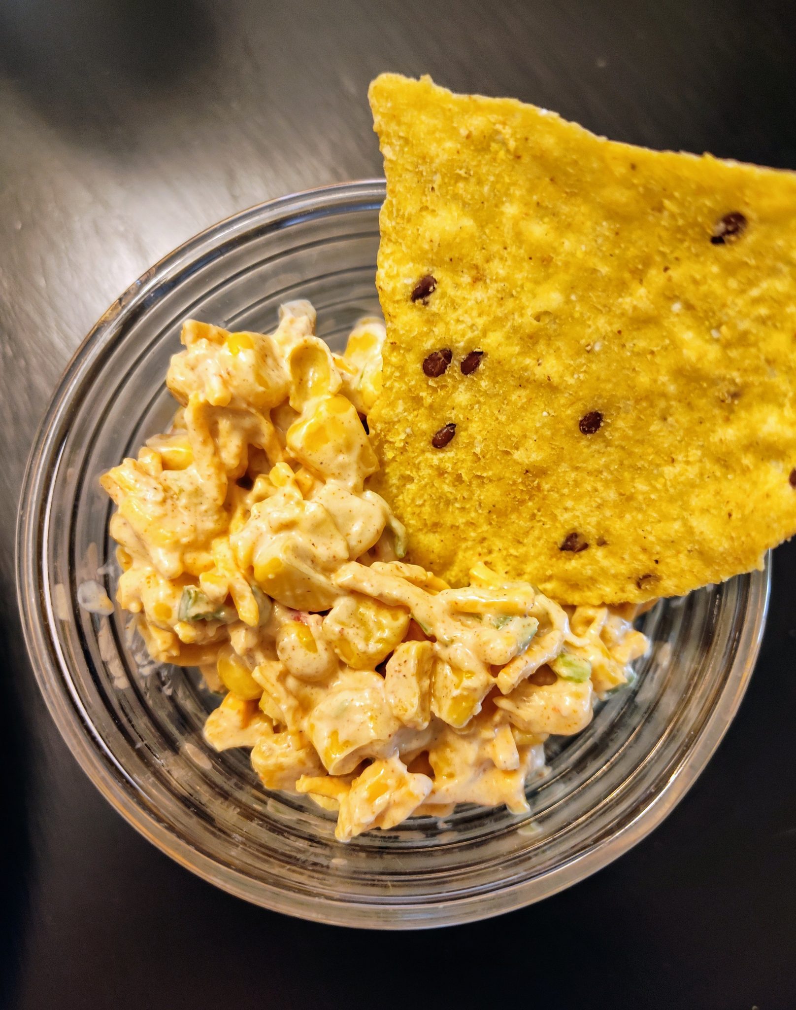 A clear bowl with creamy corn dip and a triangular tortilla chip on a dark surface.