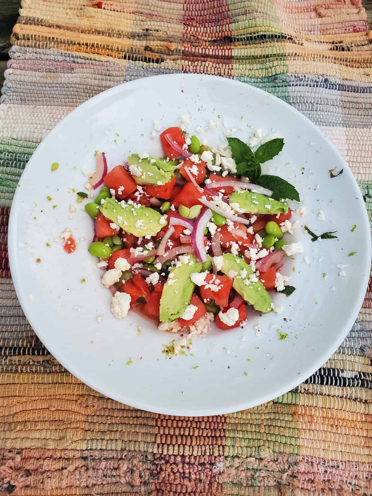 A white plate with watermelon, avocado, red onion, feta cheese, and herbs on a colorful woven placemat.