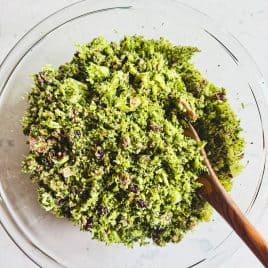 Chopped broccoli salad with bits of bacon in a glass bowl, mixed with a wooden spoon on a light surface.