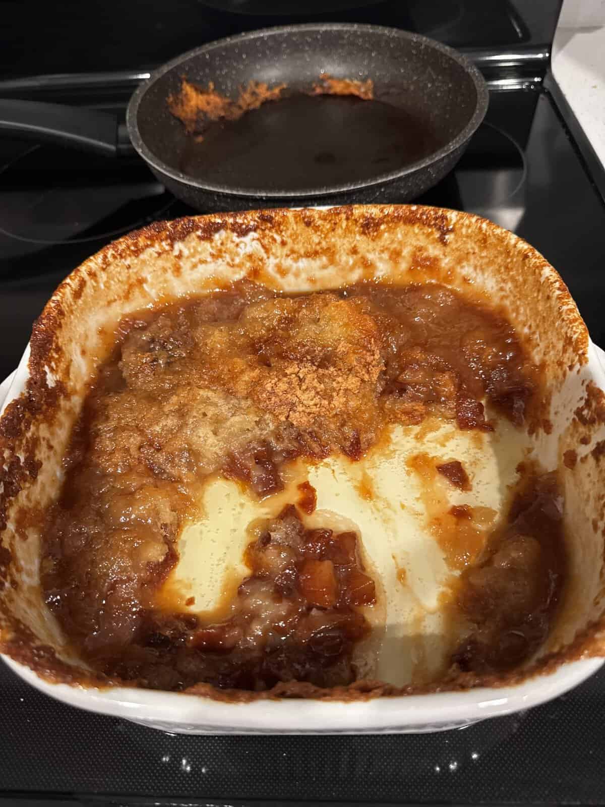 Partially eaten fruit cobbler in a white baking dish, with a used frying pan in the background on the stove.
