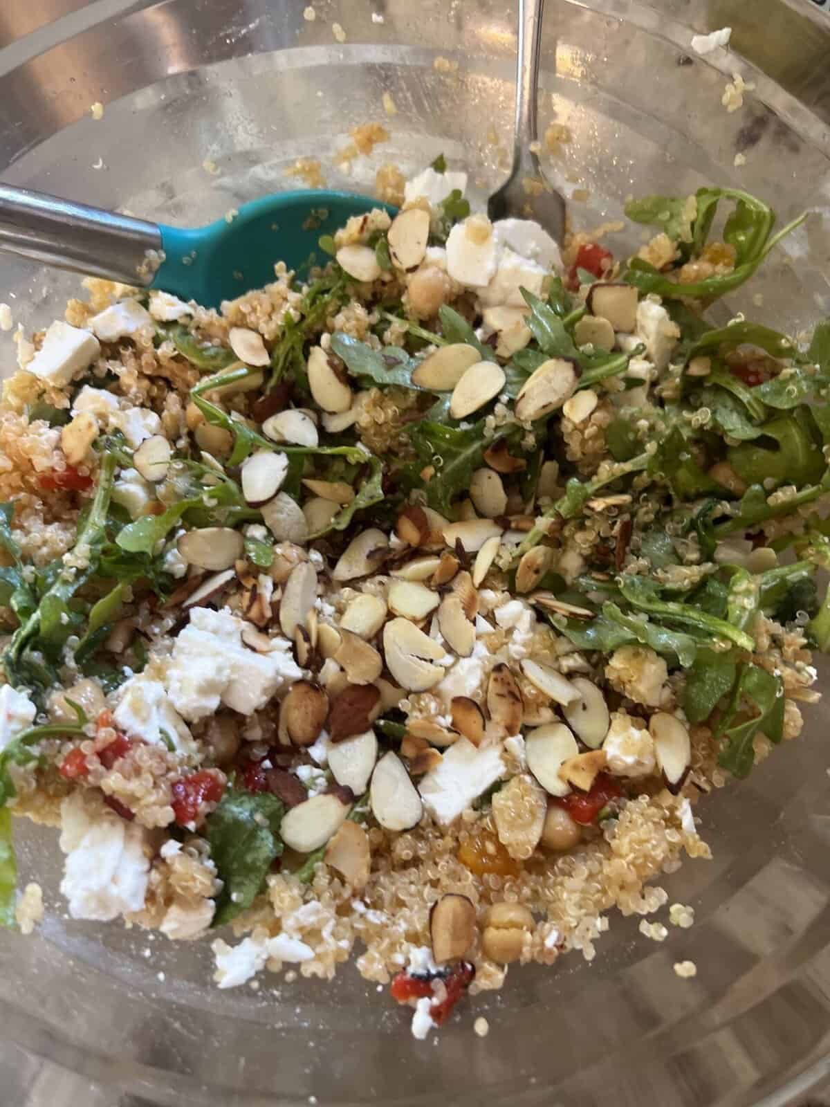 A salad with quinoa, arugula, sliced almonds, feta cheese, and chopped red peppers in a clear bowl with utensils.