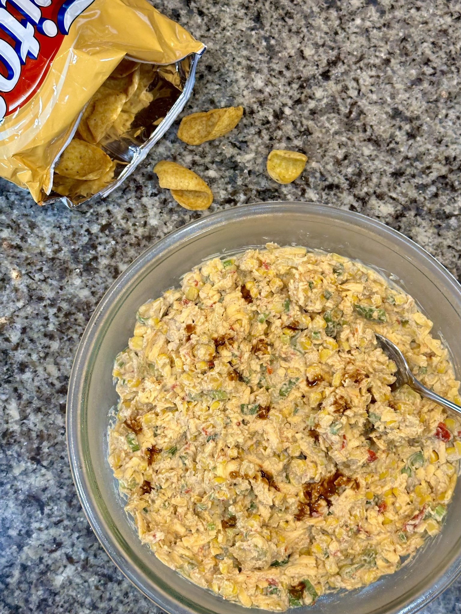 A bowl of creamy corn dip with a spoon next to an open bag of Fritos chips on a granite countertop.