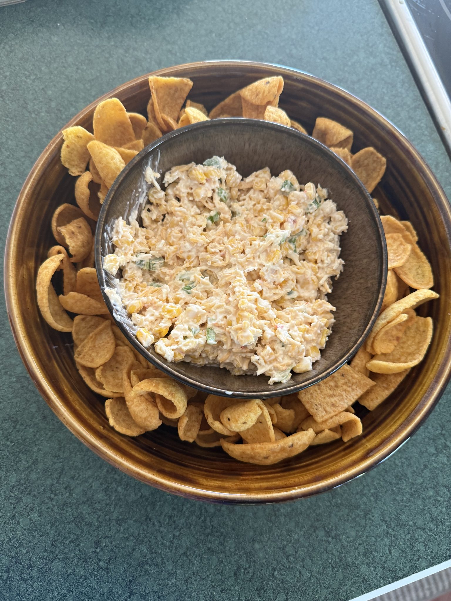 A bowl of creamy dip surrounded by corn chips on a brown plate, set on a green countertop.