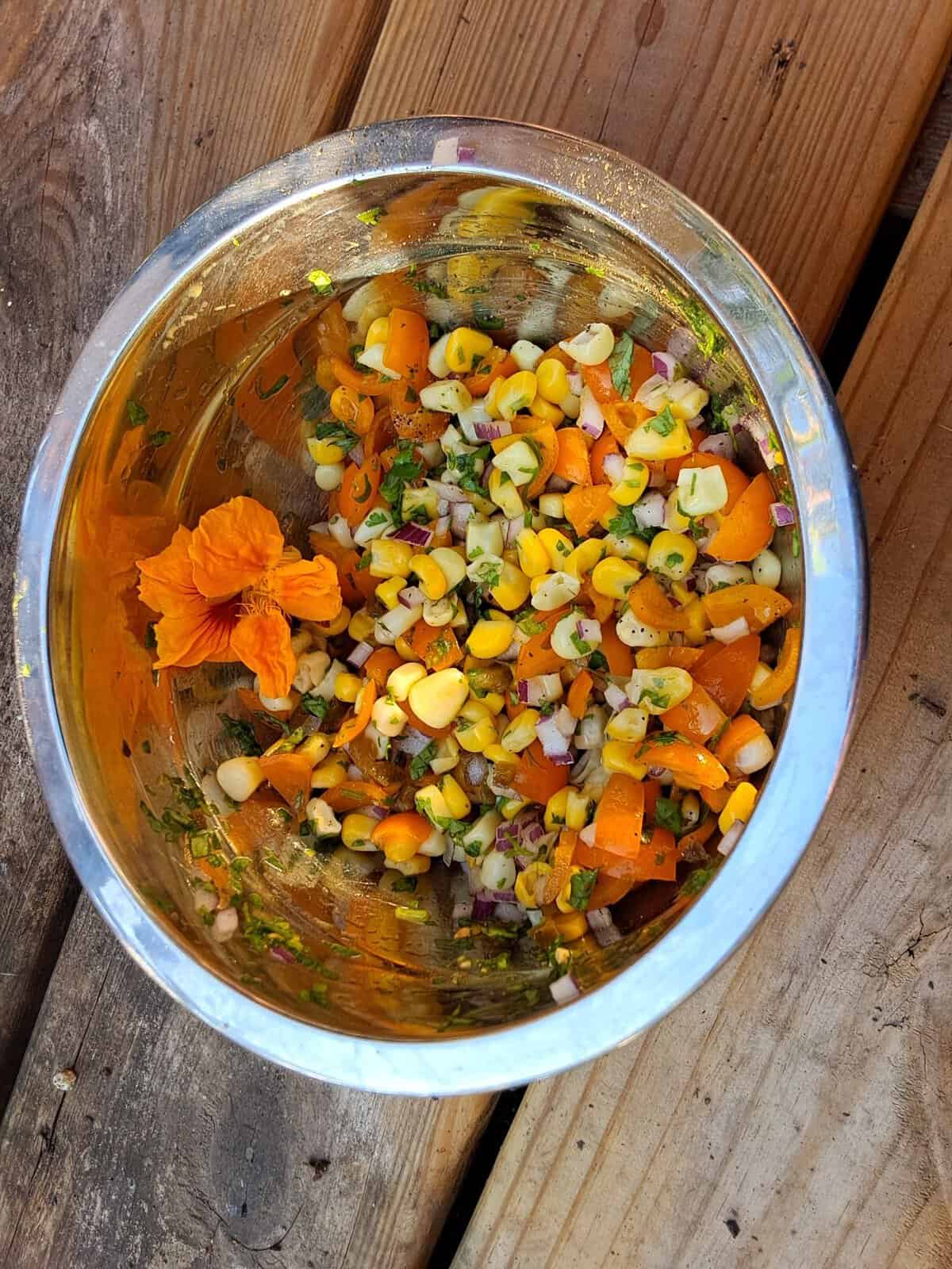 A metal bowl with corn salad, diced veggies, herbs, and an orange edible flower on a wooden surface.