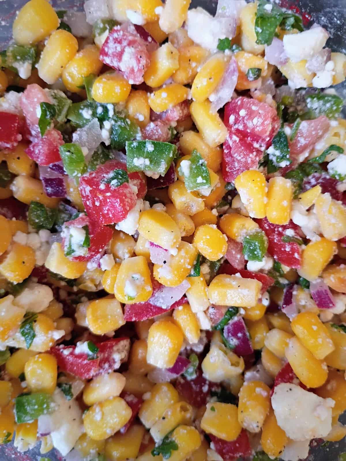 Close-up of corn salad with corn, diced tomatoes, red onion, green herbs, and crumbled cheese.