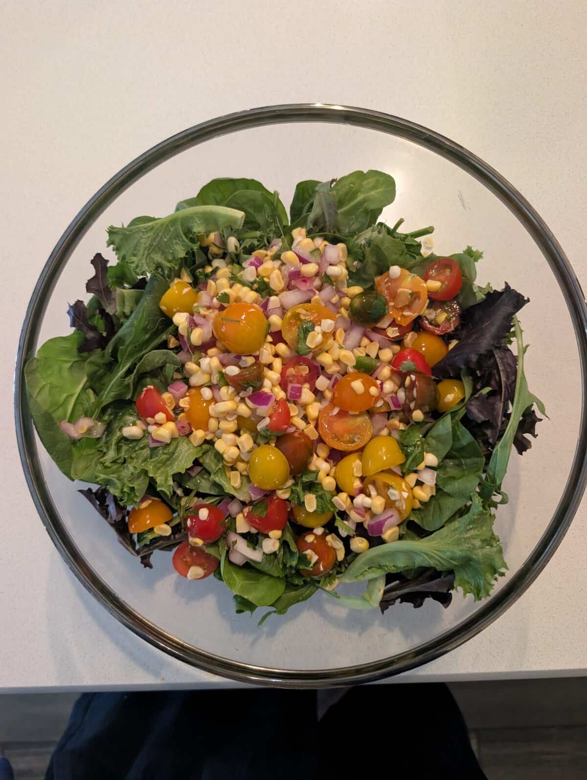 Glass bowl of mixed greens topped with cherry tomatoes, corn, red onion, and herbs on a light countertop.