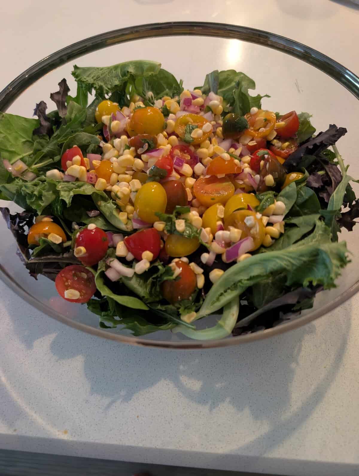 A glass bowl of salad with mixed greens, cherry tomatoes, corn, and red onion on a white countertop.