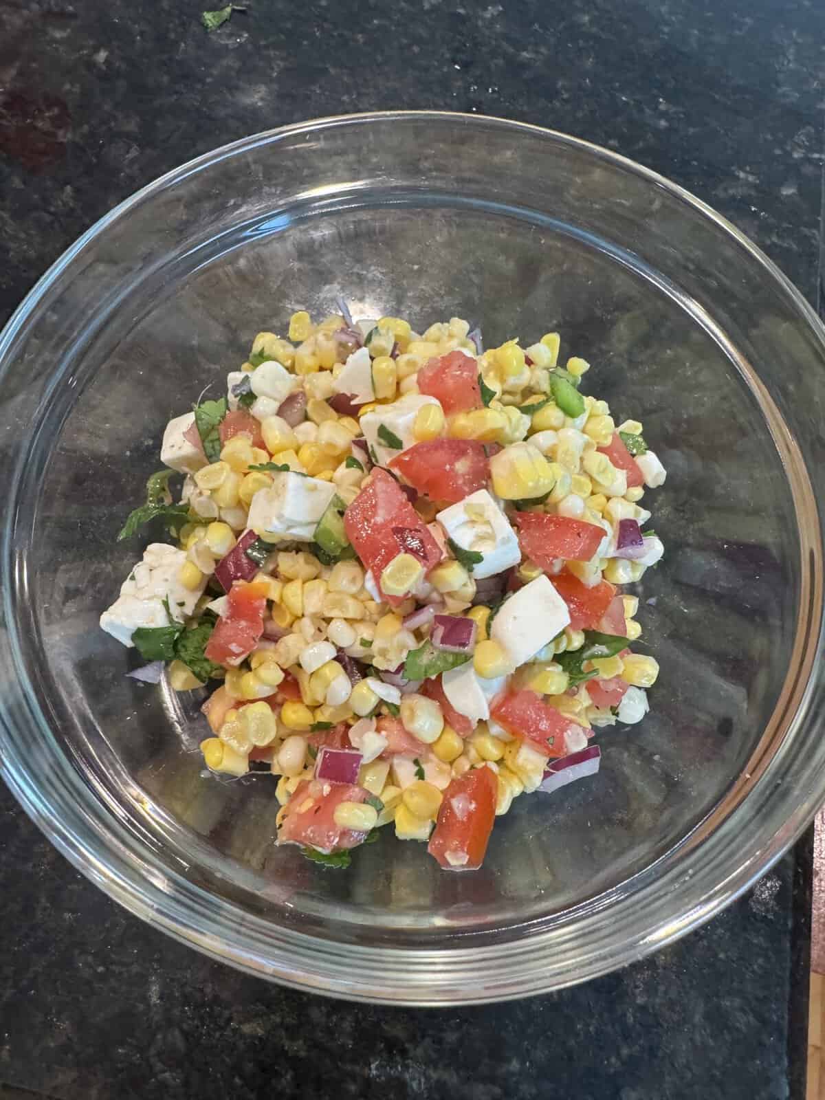 A glass bowl of corn salad with tomatoes, red onion, green herbs, and cubes of white cheese on a dark countertop.