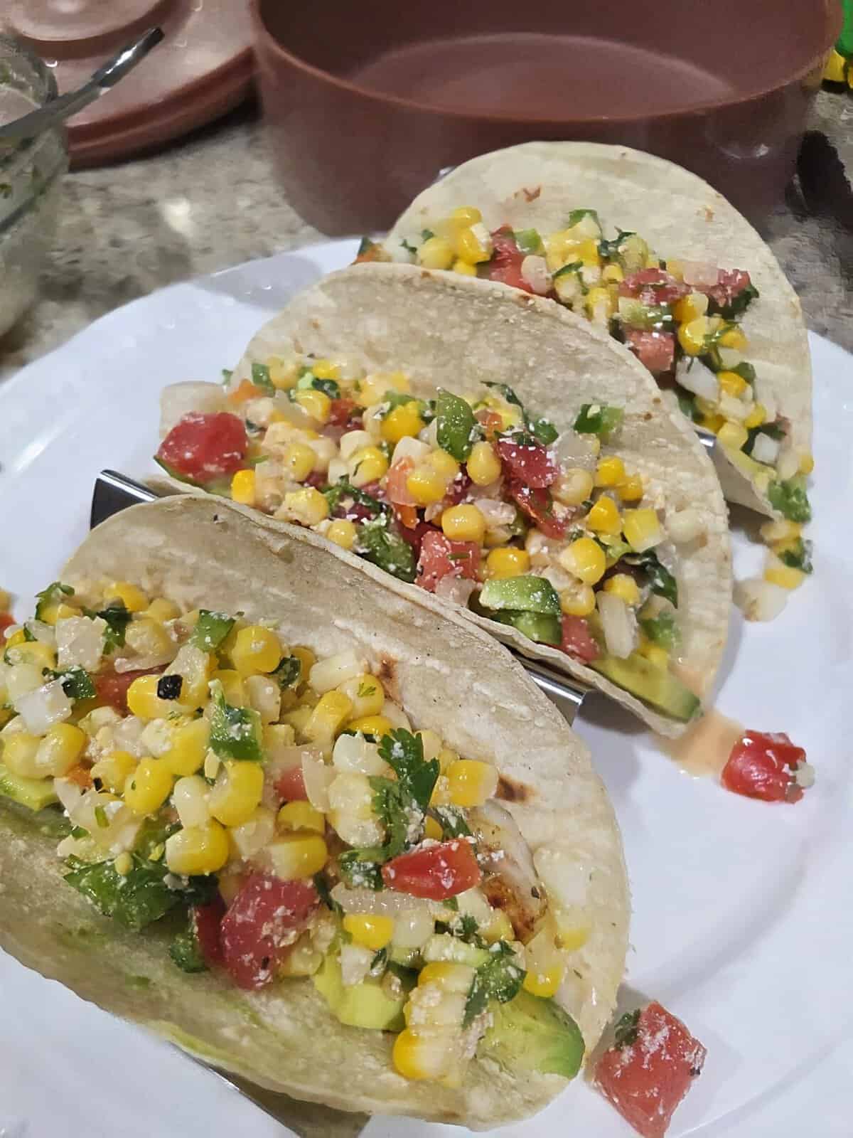 Three tacos filled with corn, tomatoes, avocado, and herbs sit on a white plate on a kitchen counter.