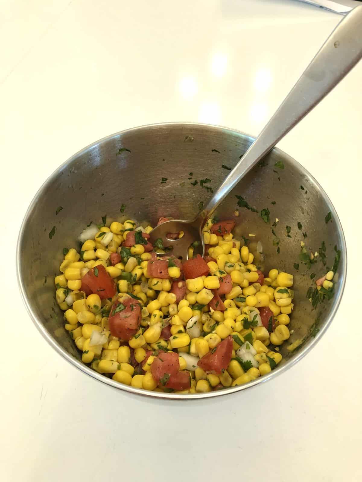 A metal bowl of corn salad with tomatoes, onions, herbs, and a spoon on a white surface.