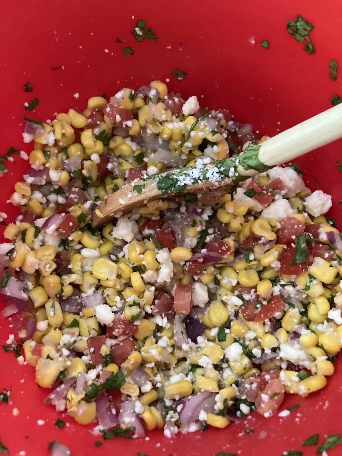 A red bowl filled with corn salad, diced onions, tomatoes, cilantro, cheese, and a wooden spoon.
