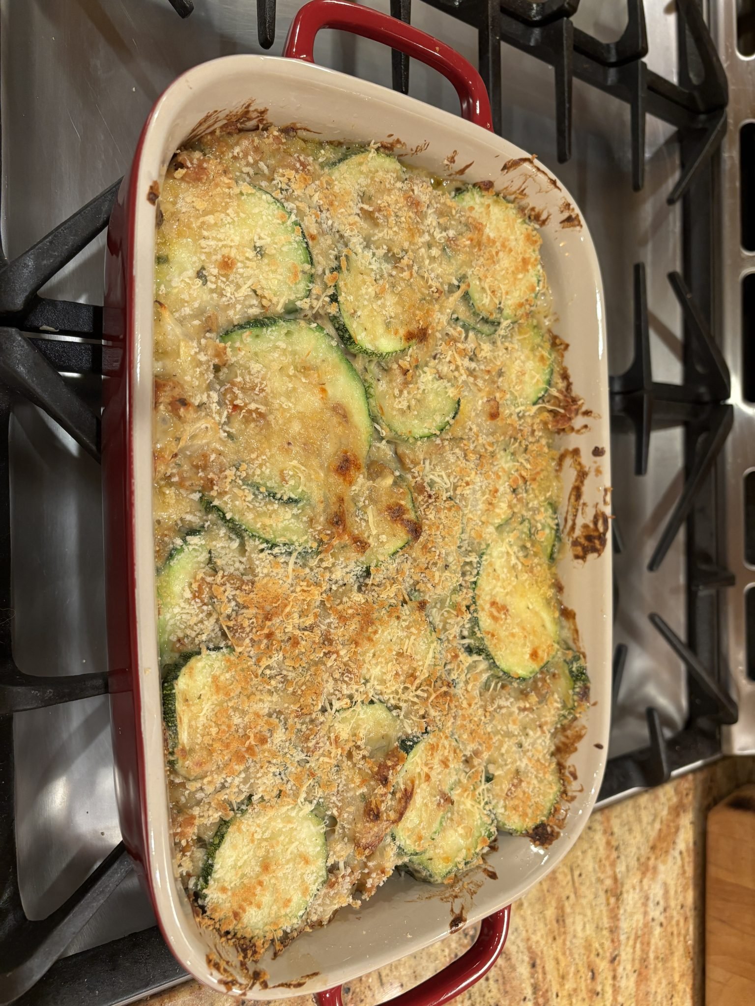 A baked casserole dish filled with golden-brown zucchini slices topped with breadcrumbs sits on a stovetop.