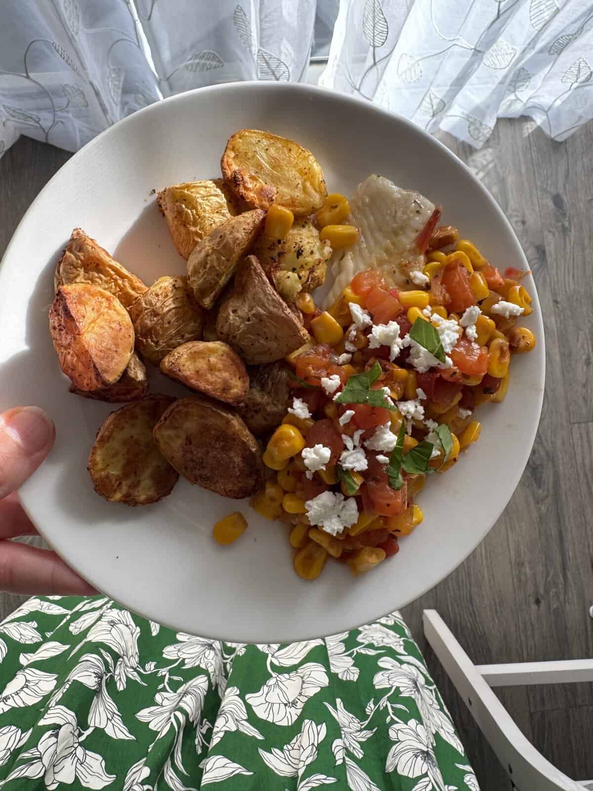 Plate with roasted potatoes, corn salsa with feta, and white fish, held above a green floral skirt.