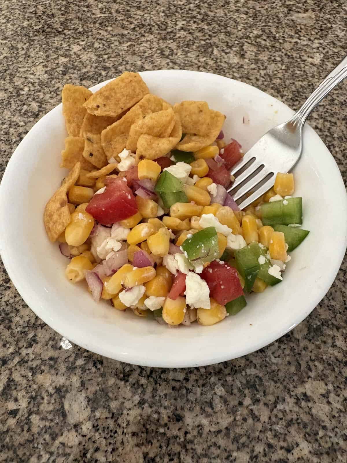 A bowl of corn salad with tomato, green pepper, feta, red onion, and corn chips on a granite countertop.