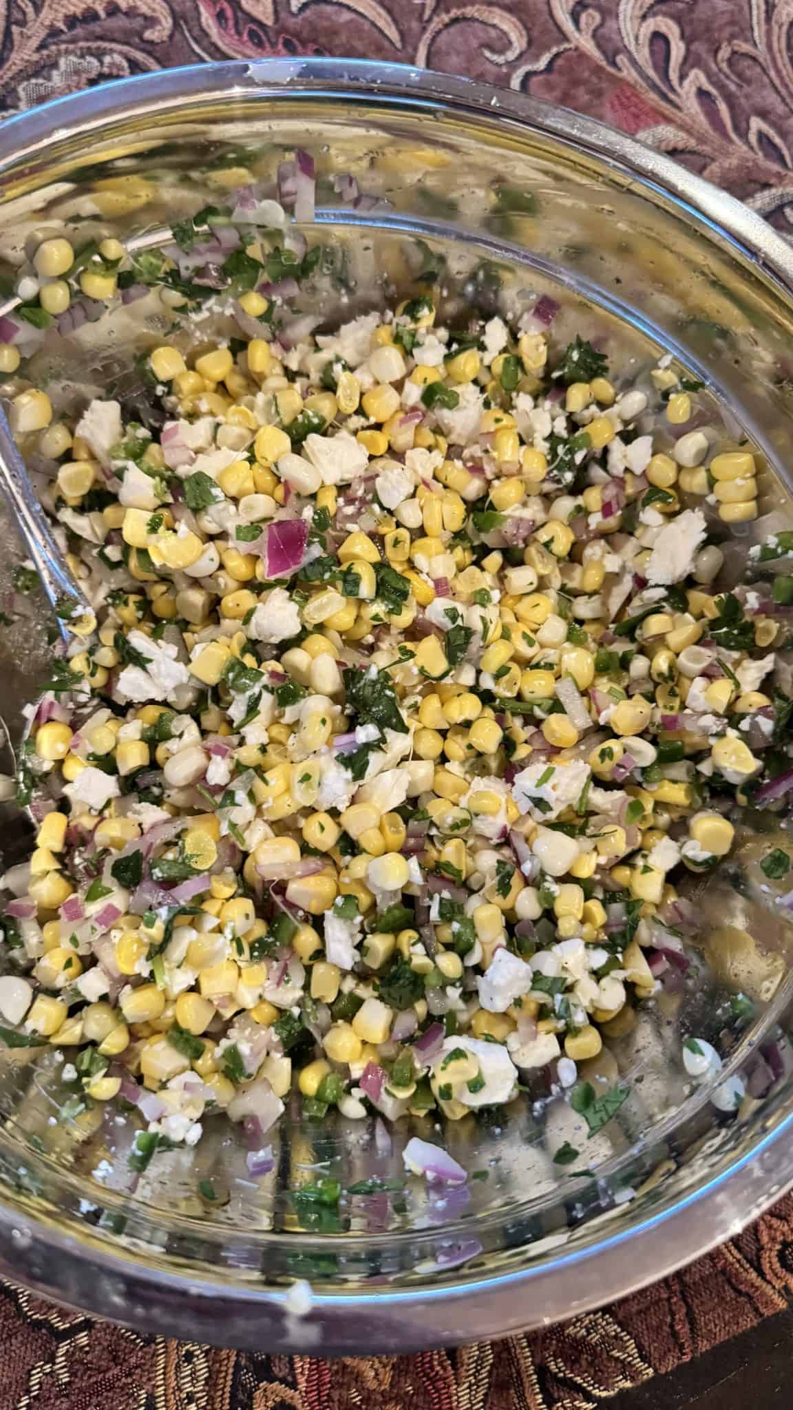 A bowl of corn salad with red onion, herbs, and cheese mixed together, set on a patterned tablecloth.