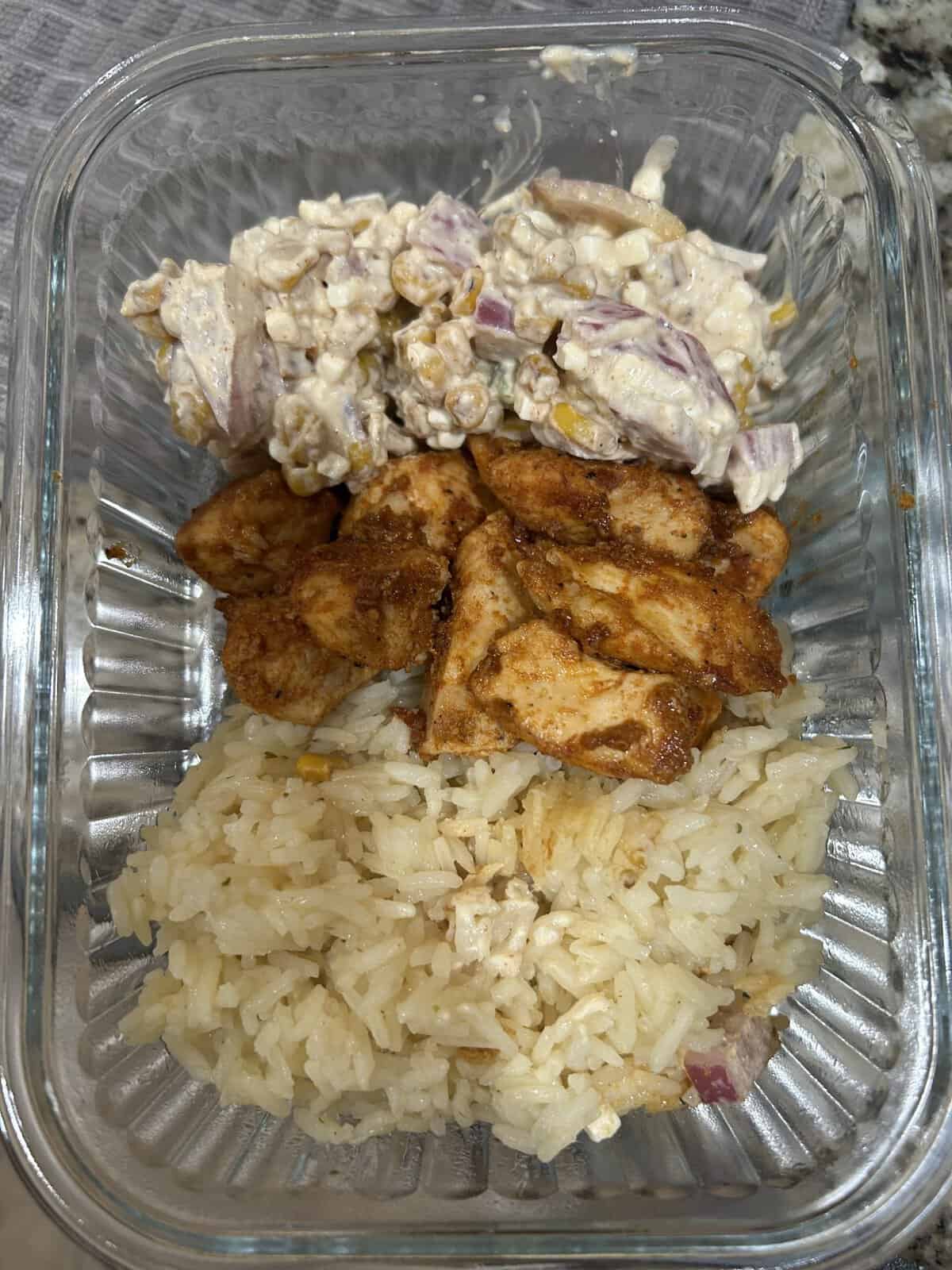 Glass container with rice, seasoned chicken pieces, and a corn and red onion salad in three separate sections.