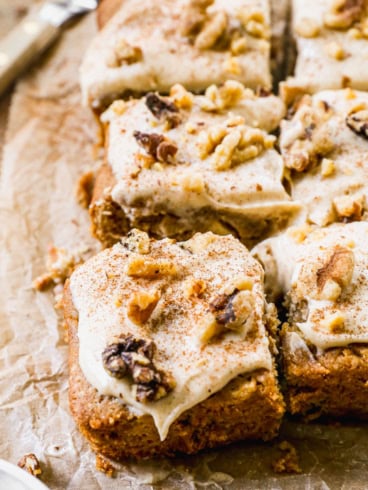 Squares of frosted cake topped with chopped walnuts and a sprinkle of cinnamon are arranged on parchment paper, with one piece slightly pulled away from the group. A knife is visible in the background.
