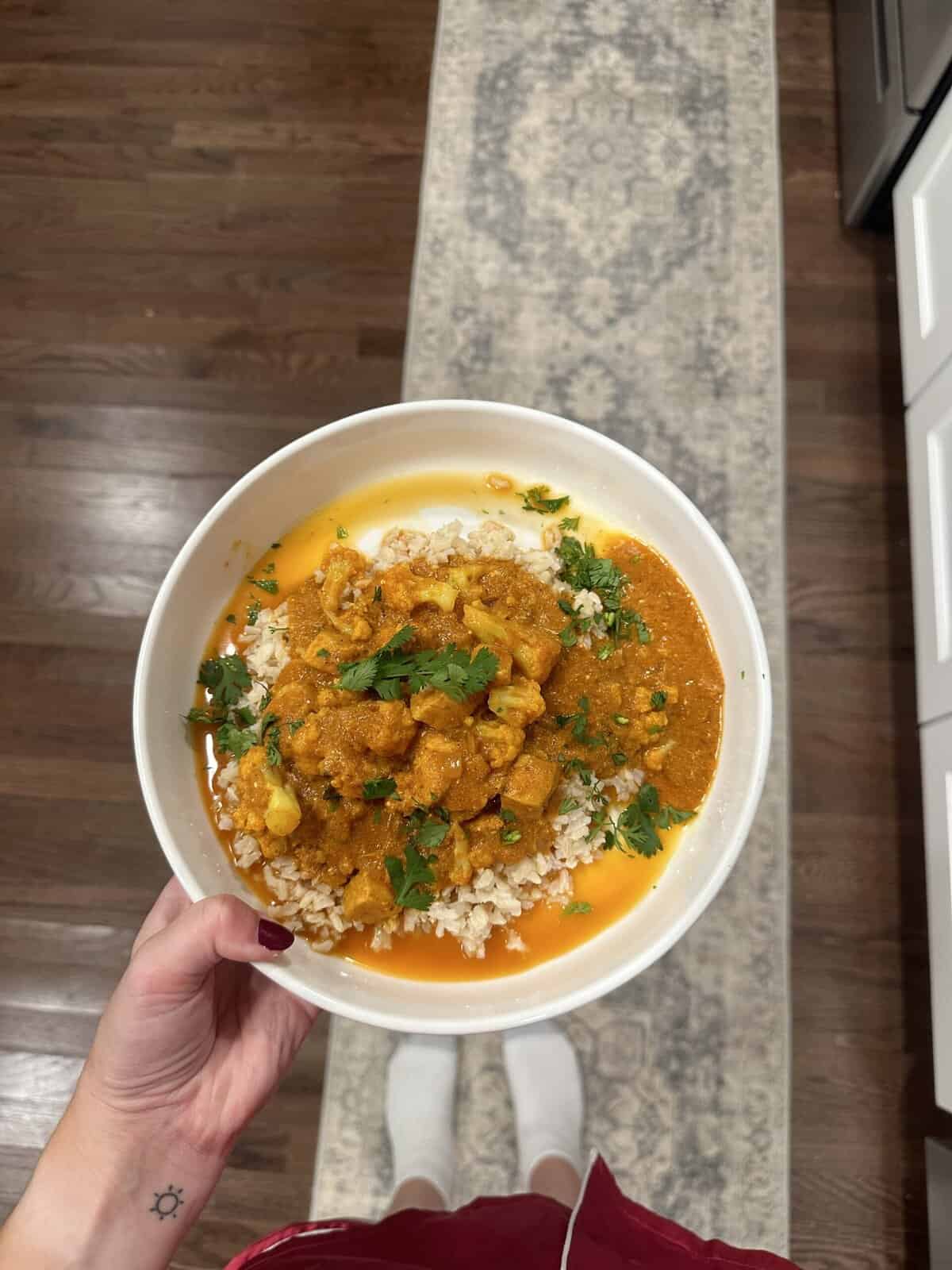 A hand holding a bowl of rice topped with orange curry and garnished with fresh herbs, above a patterned rug.