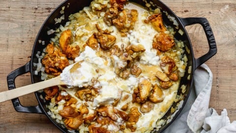 Chicken, onions, and coconut sauce cooking in a black skillet with a wooden spatula on a wooden surface.