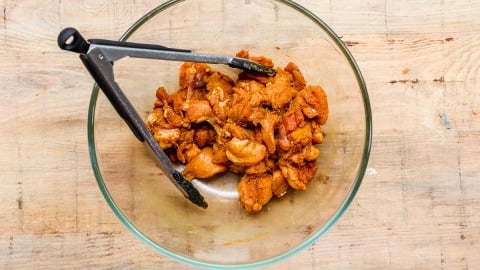 Glass bowl of marinated chicken pieces with black kitchen tongs on a light wooden surface.