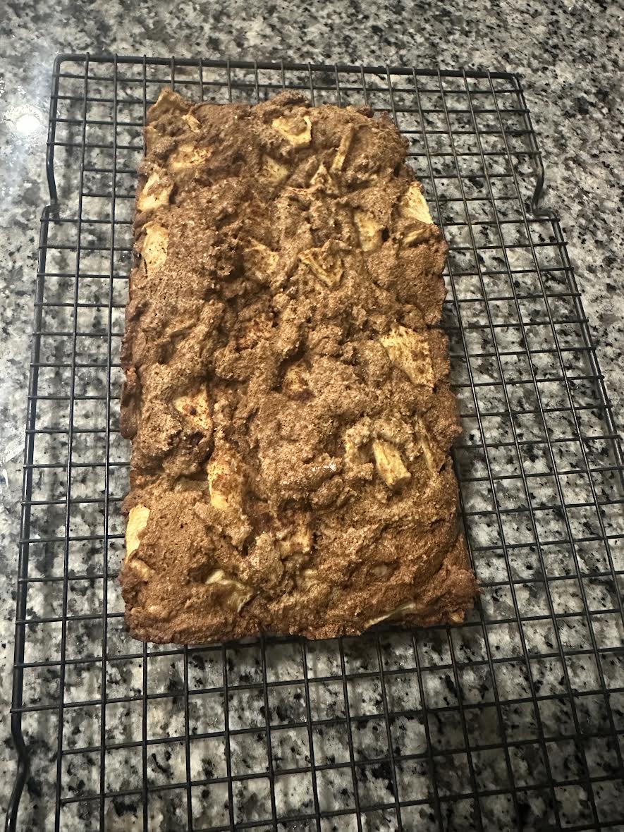A rectangular loaf of apple bread with a crumbly top cooling on a wire rack over a granite countertop.