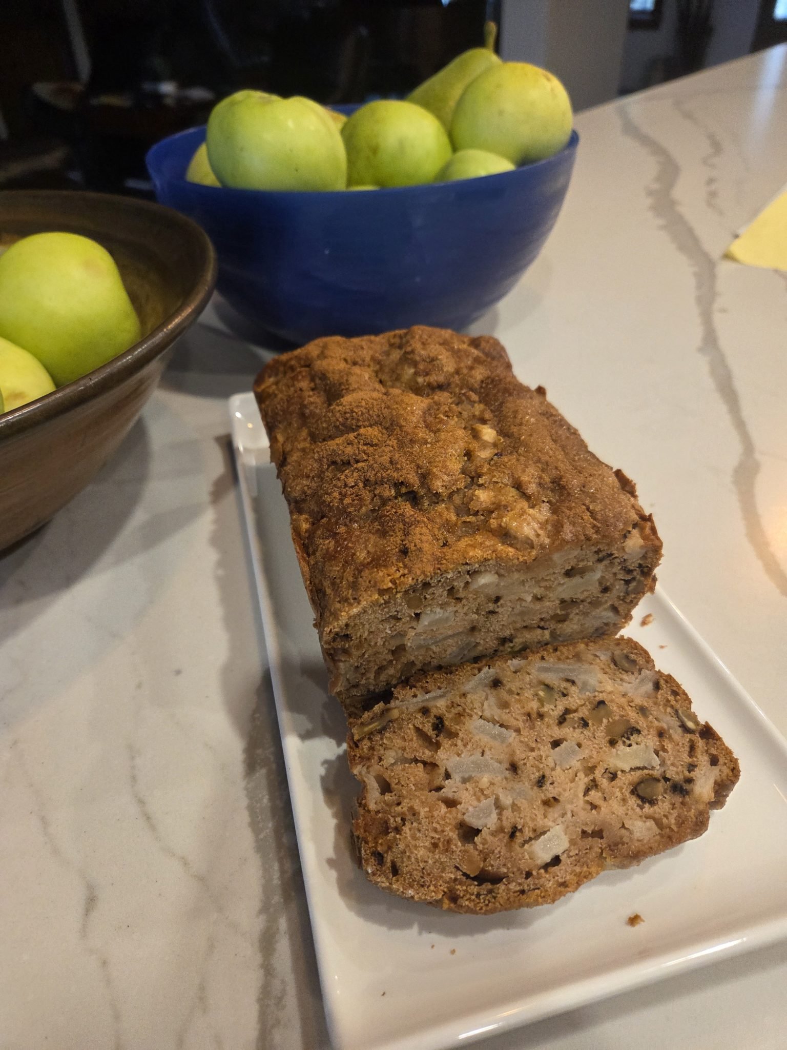 A sliced loaf of apple bread on a plate, with bowls of green apples in the background on a kitchen counter.