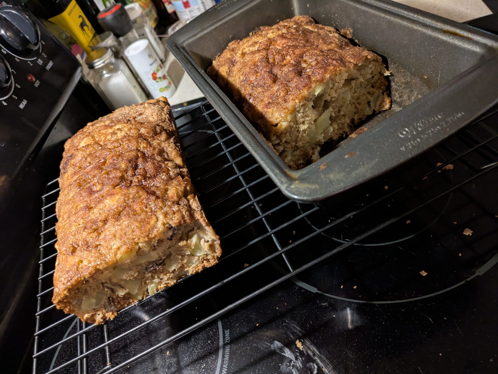 Two loaves of homemade bread, one in a pan and one cooling on a wire rack on a kitchen stovetop.