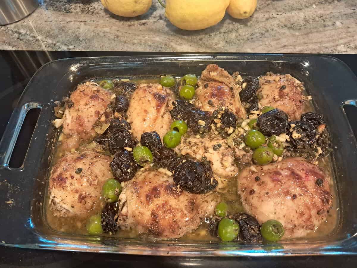 Baked chicken thighs with green olives and prunes in a glass dish on a countertop, lemons in the background.