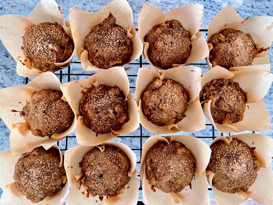 Twelve brown muffins in parchment wrappers cooling on a wire rack, viewed from above on a gray countertop.