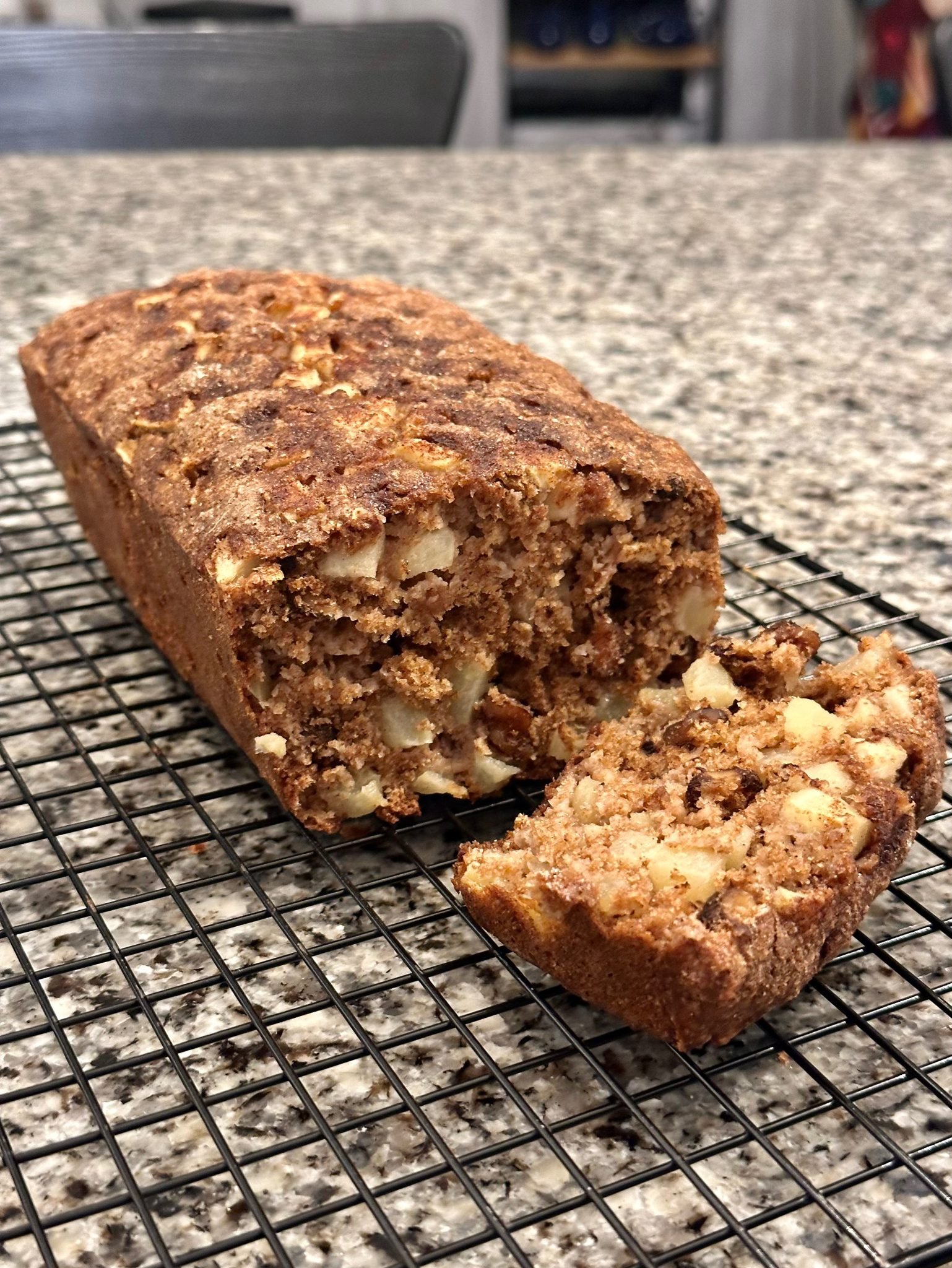 A loaf of apple bread with one slice cut, cooling on a wire rack on a granite countertop.