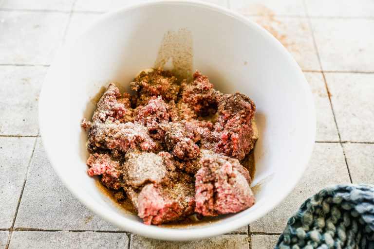 Raw ground beef with seasoning in a white bowl, sitting on a tiled countertop next to a blue cloth.