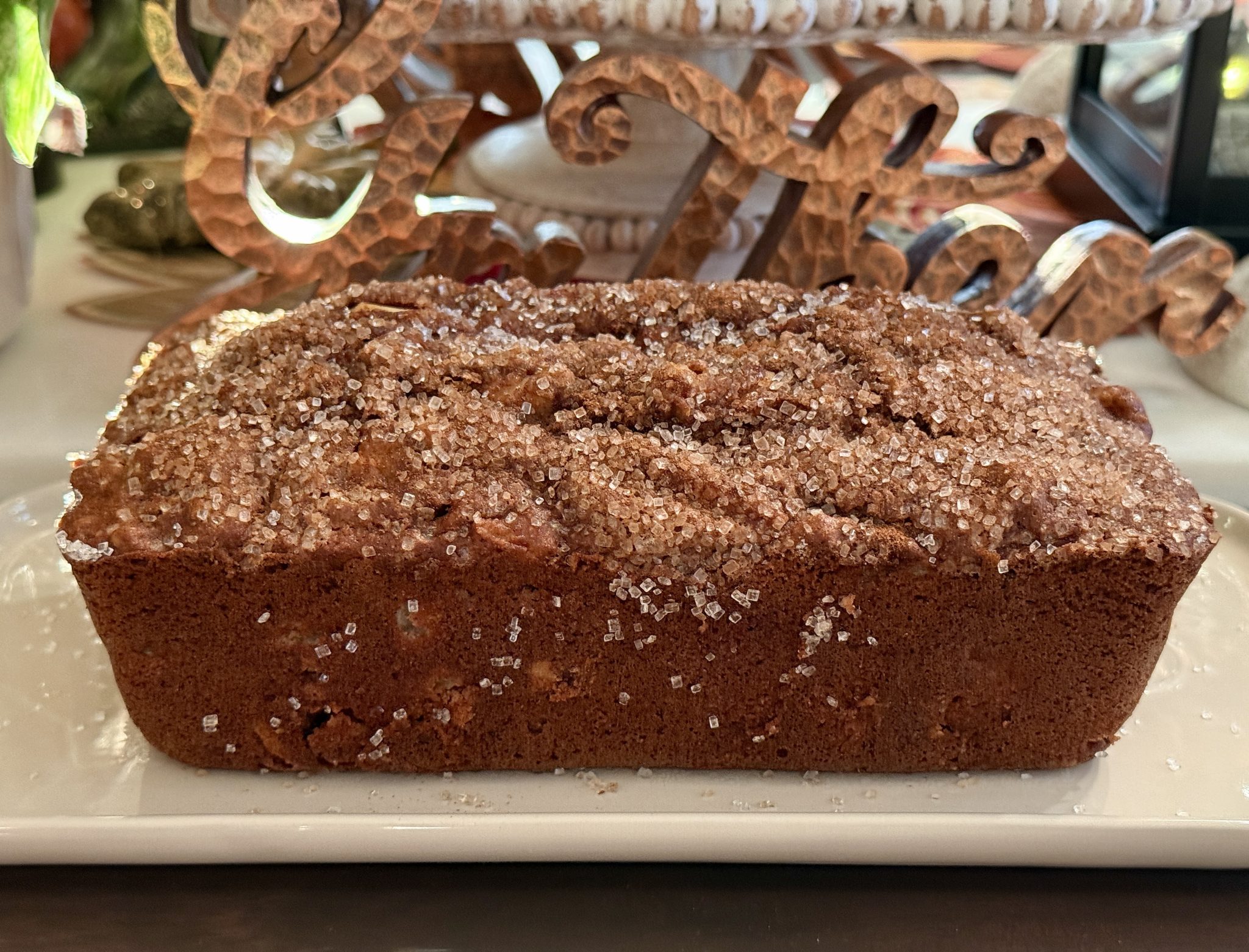 A loaf of cinnamon bread topped with coarse sugar sits on a white plate with decorative items in the background.