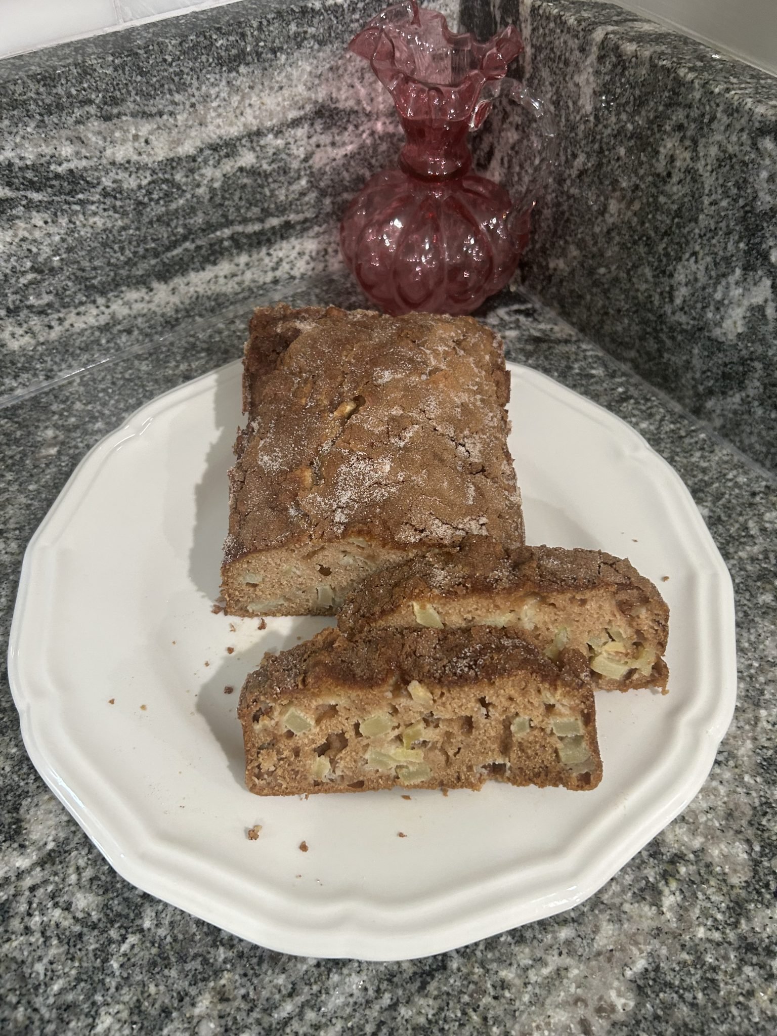 A loaf of apple bread with two slices cut sits on a white plate beside a pink glass vase on a granite counter.
