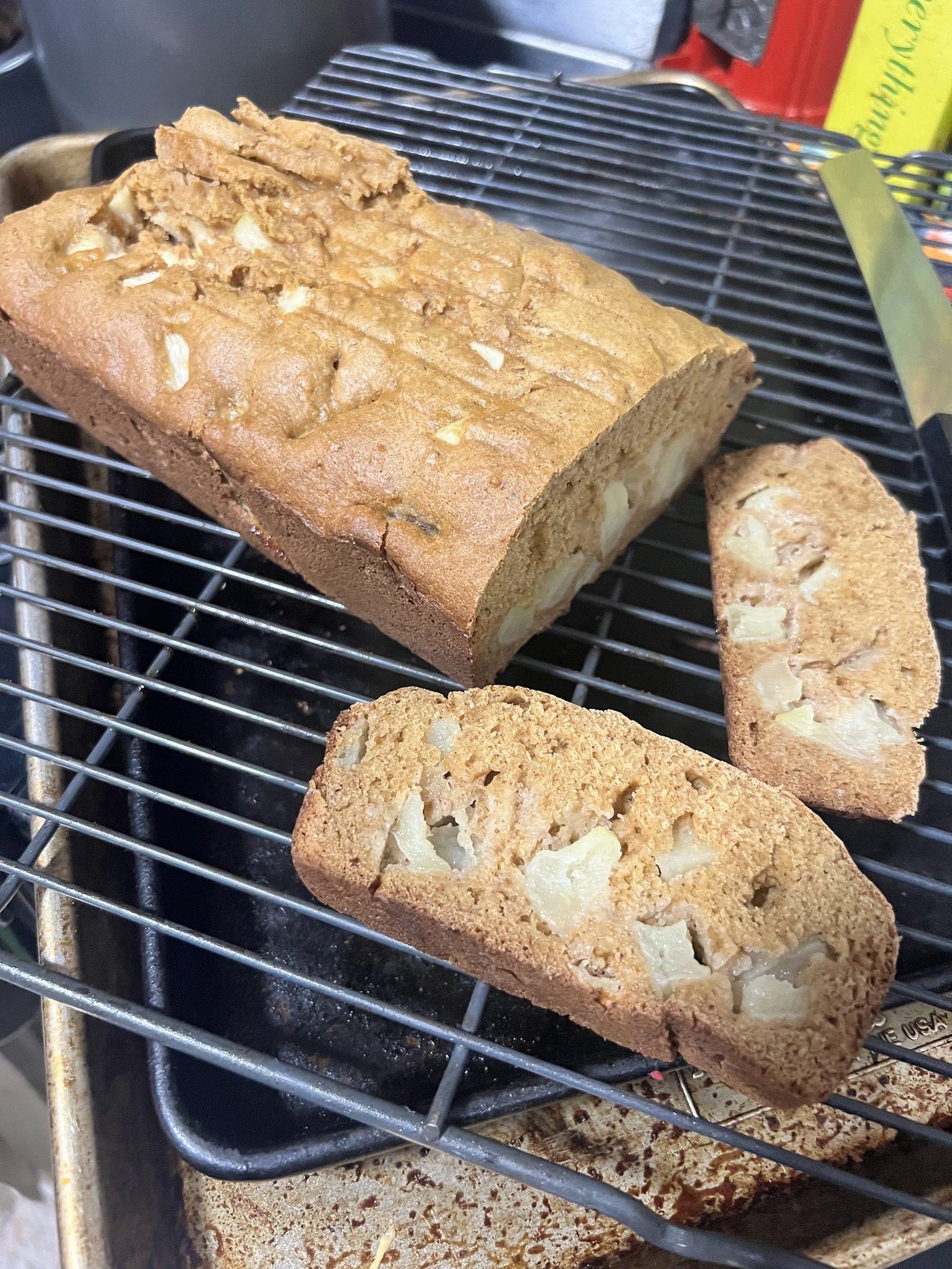 A loaf of pear bread sits on a cooling rack with two sliced pieces revealing chunks of pear inside.