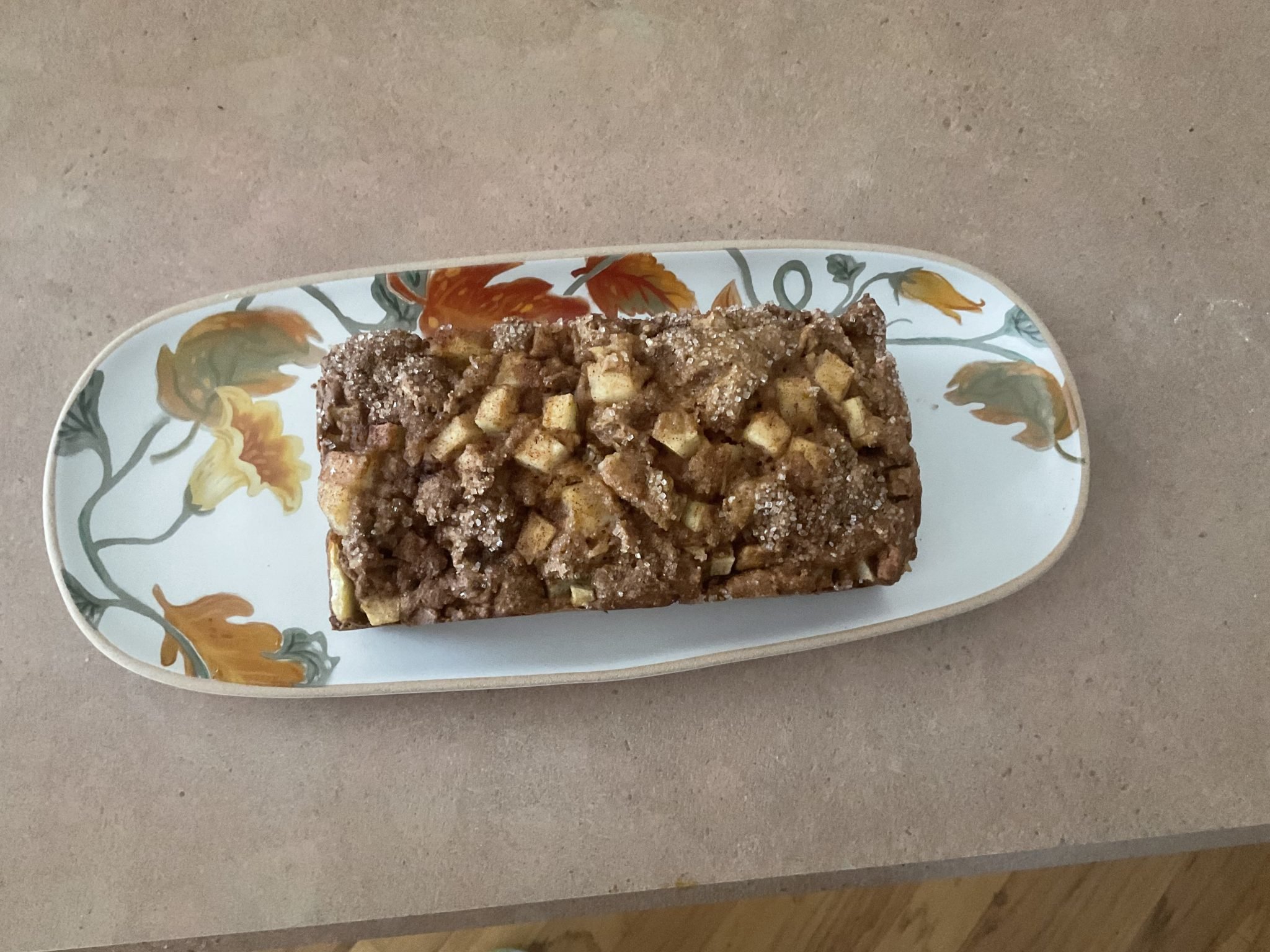 A loaf of apple cinnamon bread with crumb topping on a floral-patterned plate on a light countertop.