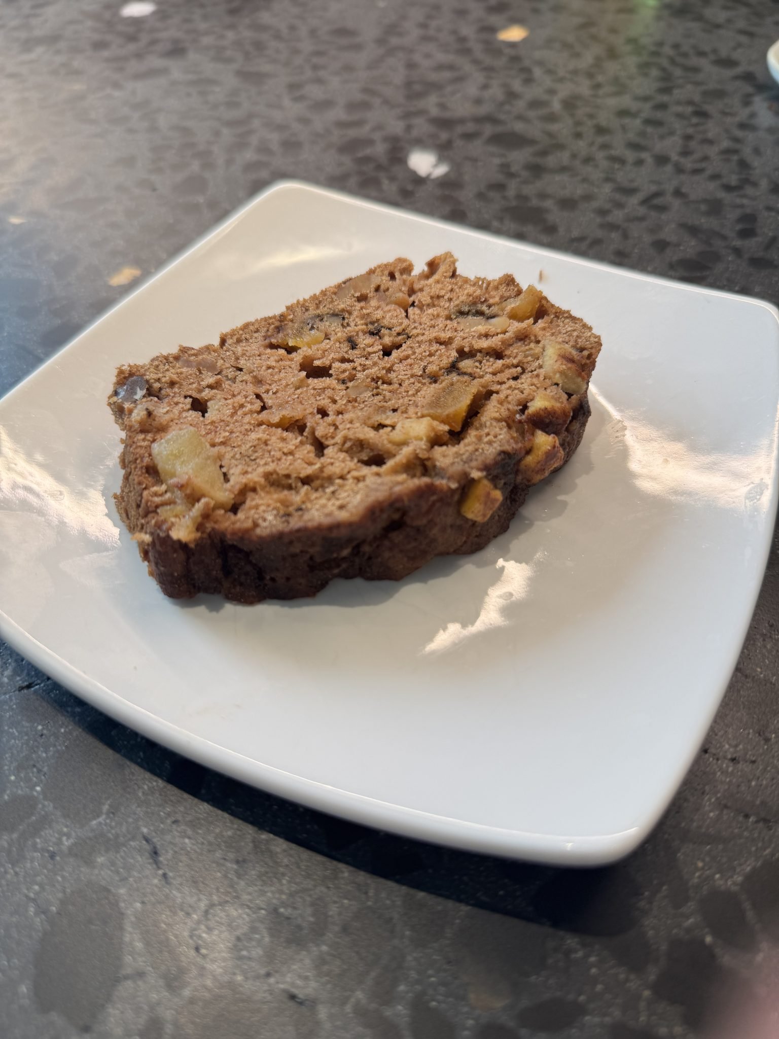 A slice of brown bread with visible fruit chunks on a white square plate, set on a dark textured surface.