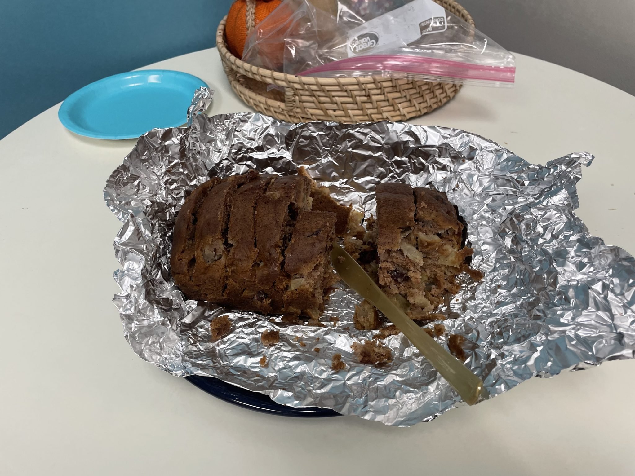 Sliced loaf cake on foil with a knife, on a table near a basket of fruit and a blue plate.