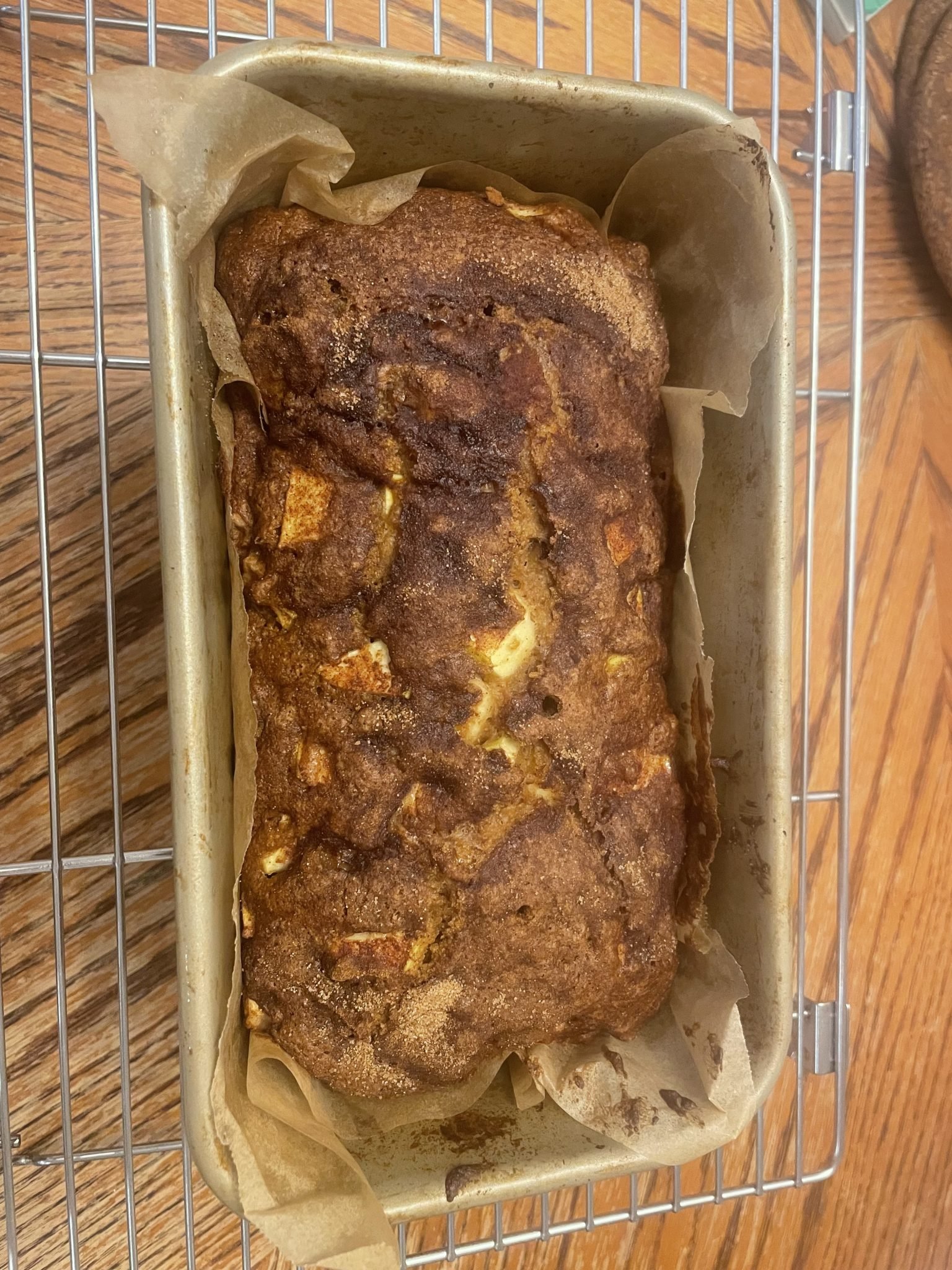 A loaf of cinnamon apple bread in a parchment-lined pan, cooling on a wire rack on a wooden table.