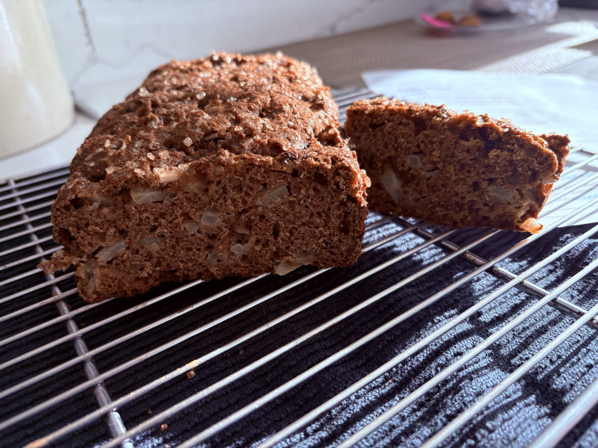 A loaf of brown bread with chopped nuts, one slice cut, resting on a cooling rack in sunlight.