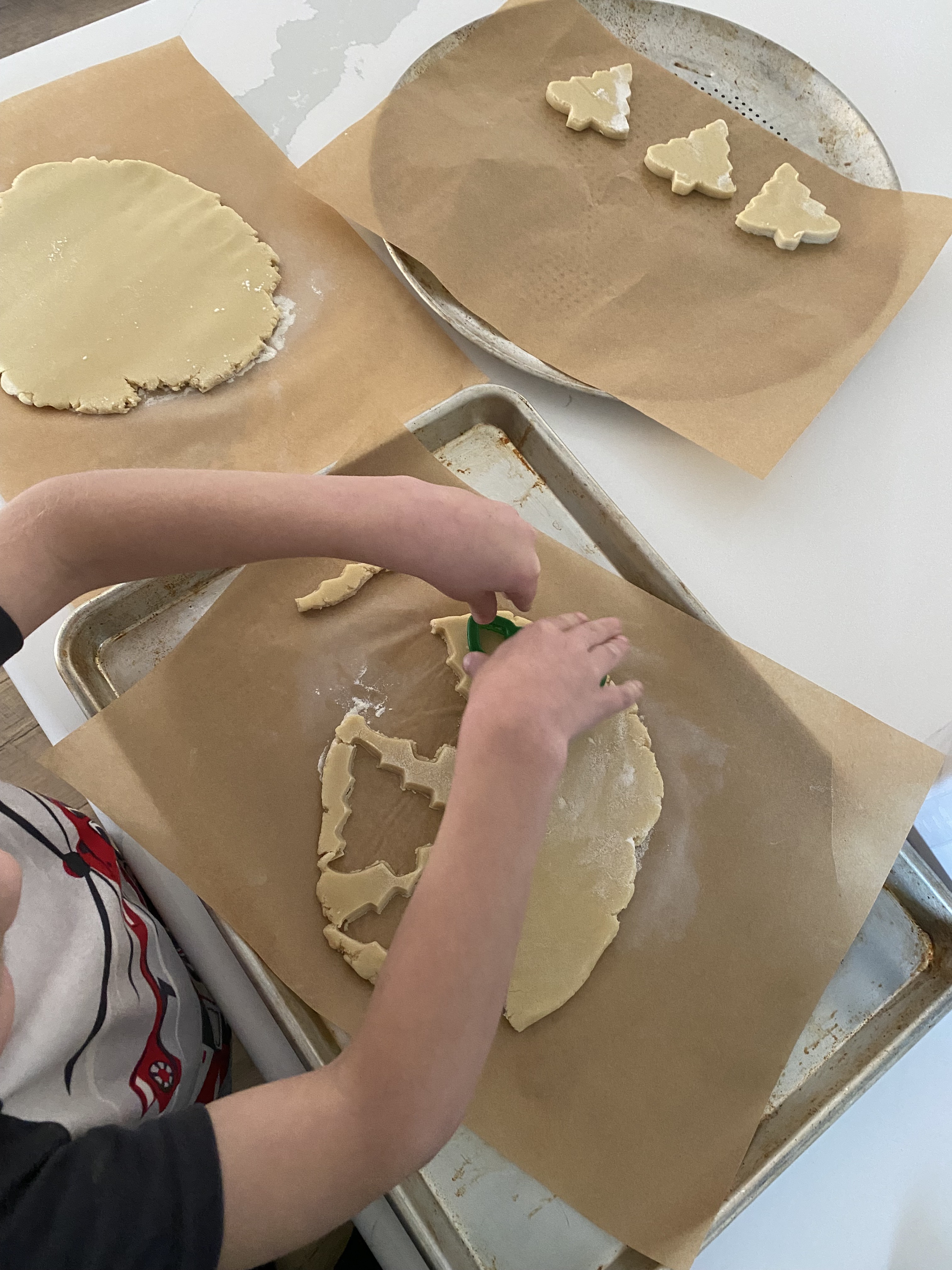 Child using a cookie cutter on dough, with tree-shaped cookies on parchment-lined baking sheets.