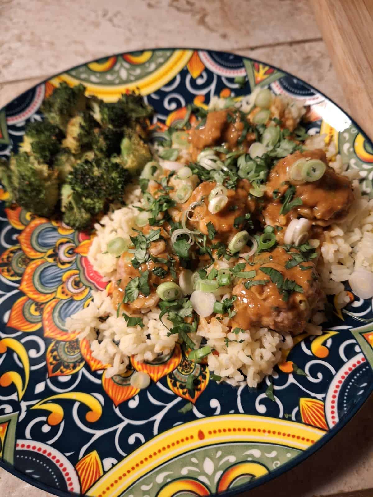 A colorful plate with rice topped with sauce, chopped green onions, and herbs, served with roasted broccoli.