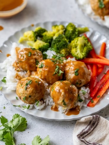 A plate with rice topped with Asian chicken meatballs, surrounded by steamed broccoli and sliced red bell peppers, garnished with chopped herbs. A beige napkin is placed beside the plate, and a small bowl of sauce is in the background.