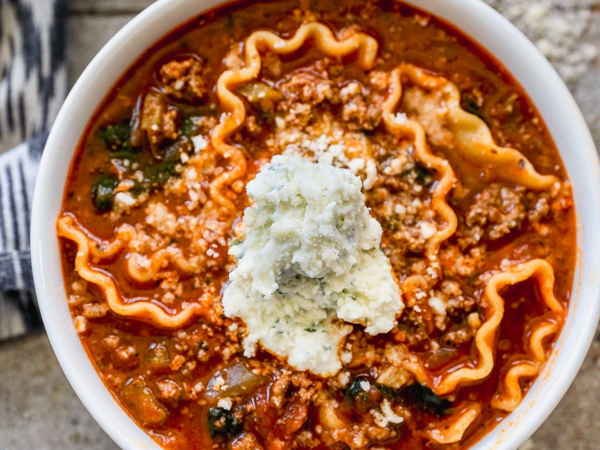 A crockpot lasagna soup, topped with a dollop of ricotta cheese, sits invitingly. The hearty dish features wavy noodles in a rich tomato-based broth infused with visible herbs and spices. Beneath the bowl, the wooden table peeks through just enough to complete the rustic setting.