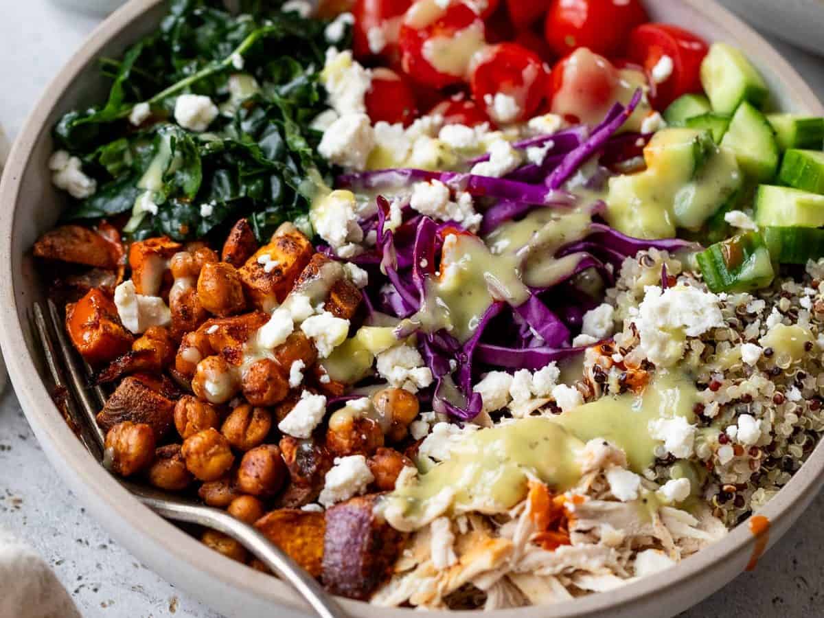 A bowl filled with roasted chickpeas, sweet potatoes, shredded chicken, quinoa, kale, cherry tomatoes, cucumber, red cabbage, and feta cheese drizzled with yellow dressing captures the essence of green goddess bowls. A small jug sits nearby over a light background.